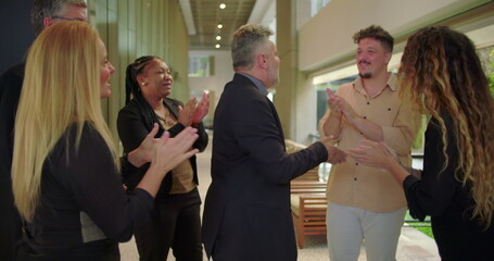 Grateful male professional smiling while receiving applause from supportive colleagues in office hallway, emotional moment of recognition and appreciation