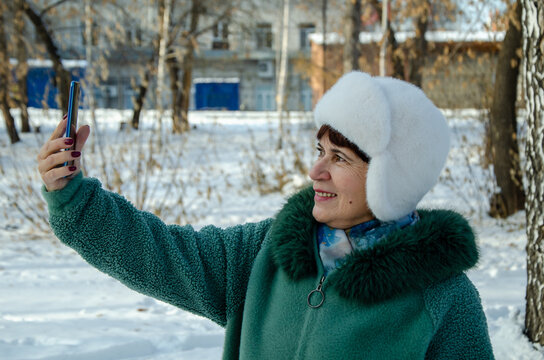 Senior woman holding your smartphone at arms length in a snowy park, photographing landscape and taking selfies