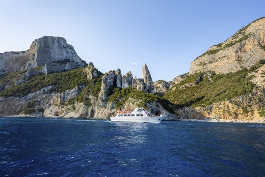 Excursion boat off picturesque rocky coast, cliffs with caves in morning light, L'Aguglia rock pin, blue sea and Cala Goloritz&eacute; beach, Golfo di Orosei, Baunei, Sardinia, Italy