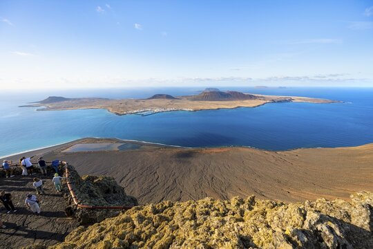 View of La Graciosa island with volcanic craters in the evening light, tourists on an observation deck at the Mirador del R&iacute;o viewpoint designed by artist C&eacute;sar Manrique, Lanzarote, Canary Islands, Spain