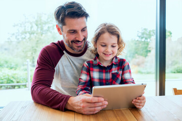 Caucasian middle aged man sitting with Caucasian child using digital tablet together at table, both smiling and looking at screen, natural daylight coming through large window