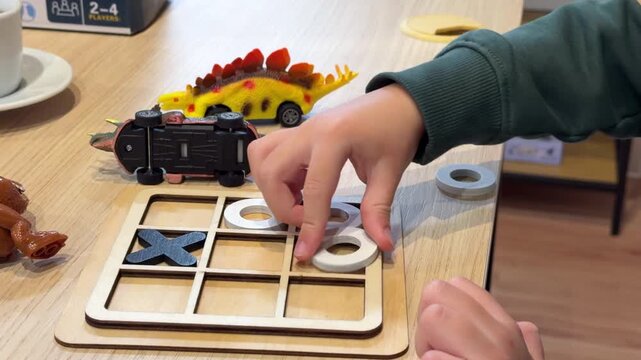 Child playing a wooden tic tac toe game at a table with toy dinosaurs and cars around. Closeup hands arranging pieces during fun educational playtime indoors.