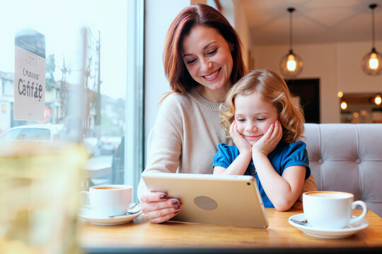Caucasian young adult woman sitting with Caucasian child girl at cafe table using digital tablet together, both smiling and looking at screen, coffee cups on table