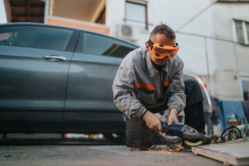 A construction worker wearing orange safety goggles and a gray work suit uses an angle grinder on a concrete surface, sparks fly as grime and dust rise.