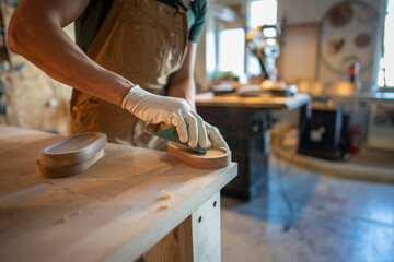 Man carpenter in apron sanding wooden coaster for water glasses with sand paper at working table, craftsman adds finishing touches for new item, working process in small business carpentry workshop.