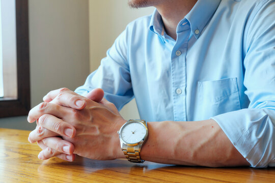 Caucasian young adult man sitting at wooden table clasping hands together wearing wristwatch visible on left wrist showing relaxed posture with natural light coming from window - Powered by Adobe
