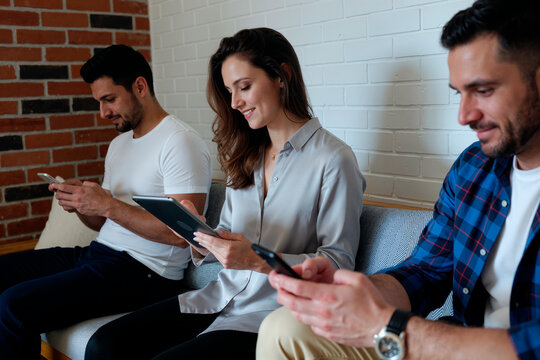 Three young adult Caucasian people sitting on sofa using digital devices, two men holding smartphones while woman in center using tablet, all focused on screens