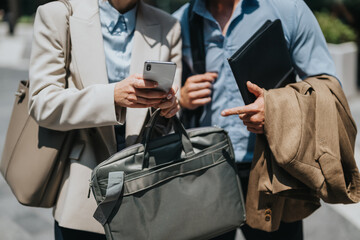 Two colleagues stand outdoors, one scrolling a phone while the other holds a folder and jacket....