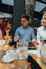 Three professionals in smart casual attire talk and gesture across a wooden cafe table. Glasses and tablets indicate a collaborative business discussion in a bright, modern cafe setting.