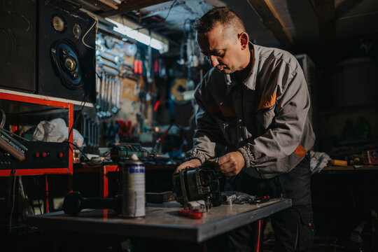 A skilled mechanic in a dim workshop repairs a machine at a metal workbench. Tools, spare parts, and equipment surround him as he concentrates on the task.