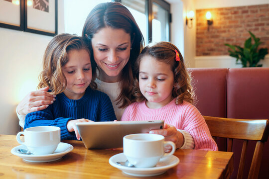 Caucasian young adult woman sitting with two Caucasian children using digital tablet together at cafe table, smiling and engaging with technology, coffee cups on wooden surface - Powered by Adobe