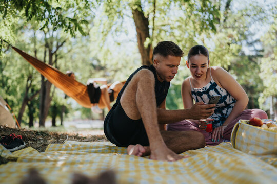 A man and woman sit on a checkered blanket in a park, sharing a moment while browsing a smartphone. A picnic basket and fruit add to the relaxed outdoor scene with a hammock in the background.