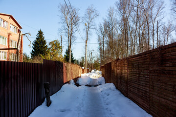Narrow snow-covered path between high fences leading towards bright sunlit distance, winter backyard scene