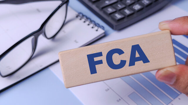 FCA (Financial Conduct Authority) Concept: Wooden Block with Abbreviation Held Over Financial Chart, Calculator, and Notebook