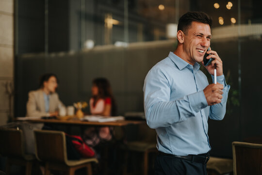 A smiling man talks on his phone in a bright, contemporary office cafe while coworkers meet at a table in the background, capturing a lively, collaborative business environment. - Powered by Adobe