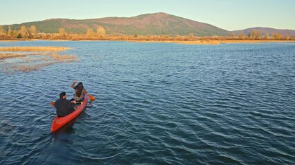 A couple paddles a red canoe on a serene lake, surrounded by autumnal trees and distant mountains. The scene captures a peaceful weekend activity, emphasizing connection, adventure.