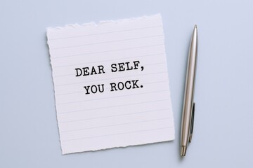 Inspirational message on paper with pen: Dear self, you rock on blue background, promoting positive self-talk.