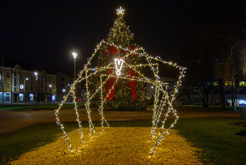 Illuminated holiday light tunnel installation leading toward a decorated Christmas tree with red bows and glowing ornaments in a city square at night.