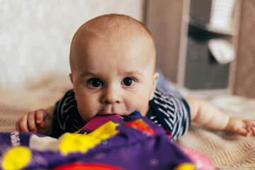 A cute baby sits on a soft chevron-patterned blanket, holding a bright yellow rattle toy. The child, dressed in a pink long-sleeve shirt and dark overalls, looks directly at the camera with a curious 