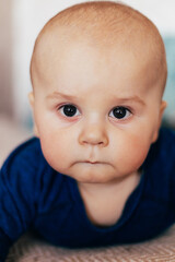 A cute baby lying on their stomach on a bed, looking forward with a curious expression. Warm natural light and a cozy home environment create a tender and intimate family scene.