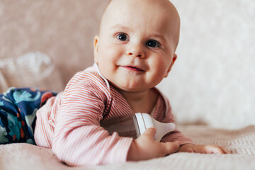 The photo shows a smiling baby lying on a soft blanket. The child is wearing a pink-and-white striped outfit and holding a baby bottle. The warm, light background gives the scene a cozy, homey feeling
