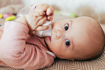 The photo shows a smiling baby lying on a soft blanket. The child is wearing a pink-and-white striped outfit and holding a baby bottle. The warm, light background gives the scene a cozy, homey feeling