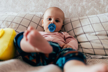 A baby is lying on a bed, leaning against checkered pillows and looking calmly at the camera. The child has a blue pacifier and is dressed in a striped shirt and colorful pants. The relaxed home 
