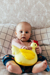 A cute baby sits on a soft chevron-patterned blanket, holding a bright yellow rattle toy. The child, dressed in a pink long-sleeve shirt and dark overalls, looks directly at the camera with a curious 