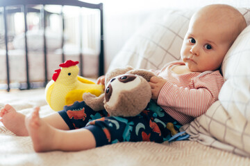 A cute baby sits on a soft chevron-patterned blanket, holding a bright yellow rattle toy. The child, dressed in a pink long-sleeve shirt and dark overalls, looks directly at the camera with a curious 