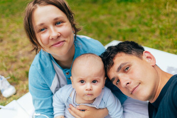 Young family taking a selfie outdoors with their one-year-old child. The parents hold the toddler close while smiling into the camera, enjoying a warm day in nature. Natural light and a relaxed atmosp