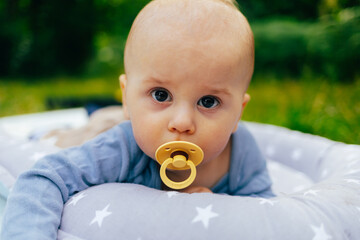 The photo shows a baby lying on a soft star-patterned cushion outdoors. The child is looking directly at the camera while sucking on a pacifier. The warm sunlight and green background create a peacefu