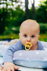 The photo shows a baby lying on a soft star-patterned cushion outdoors. The child is looking directly at the camera while sucking on a pacifier. The warm sunlight and green background create a peacefu