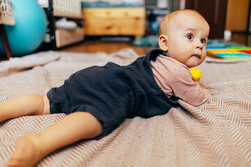 A cute baby lying on their stomach on a bed, looking forward with a curious expression. Warm natural light and a cozy home environment create a tender and intimate family scene.