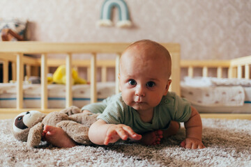 A cute baby lying on their stomach on a bed, looking forward with a curious expression. Warm natural light and a cozy home environment create a tender and intimate family scene.