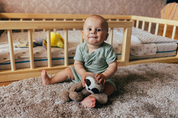 Cute baby sitting on a soft rug in a nursery next to a wooden crib. The child is holding a plush toy and looking at the camera with a gentle smile. Warm home lighting, natural tones, and a cozy atmosp