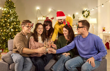 Christmas friends toasting champagne at home. Laughter fills a cozy living room as a young group in Santa hats clinks glasses at a family party. Happy holiday celebration with close friends.
