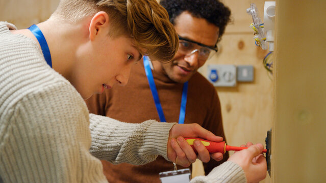 Male Tutor With Trainee Electricians In Workshop Studying For Apprenticeship At College