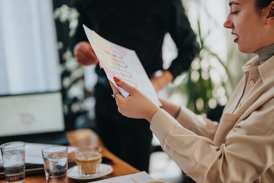 Two young coworkers sit at a table, one holding a document while another stands in the background. They discuss ideas over coffee, creating moments of teamwork and planning in a modern office.