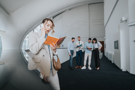 A stylish woman in a beige blazer speaks on a mobile phone while reading an orange notebook. In the background, colleagues consult tablets and papers in a bright, contemporary office corridor.