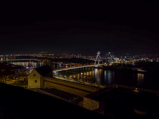 Fototapeta premium Illuminated SNP Bridge with its UFO tower rises over the Danube River at night, showcasing Bratislava’s modern cityscape and waterfront lights.