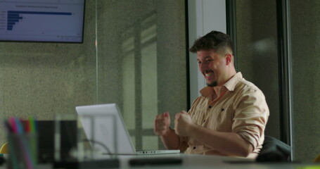 Fototapeta premium Happy young man celebrates success alone at laptop with double fist pump during solo office work session