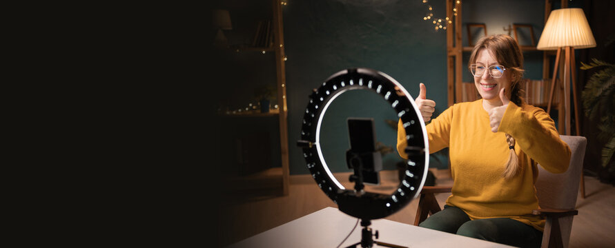 Female blogger recording video using smartphone making live selfie, social media content live video streaming making thumbs-up like gesture late evening at home office. social media