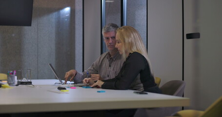 Male and female coworkers in discussion at conference table reviewing information on laptop, collaborating during late office hours in modern workplace