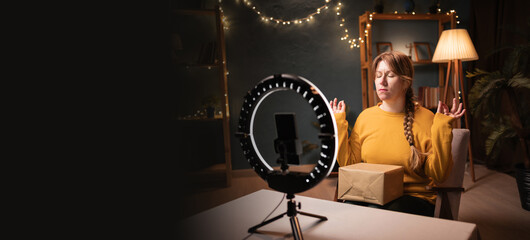 Female blogger unpacking box recording video using smartphone, ring light with tripod meditating in yoga position late evening at home office. Concept of online shopping and social media