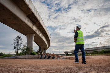 A male civil engineer or site supervisor, wearing a white hard hat, and a neon vest, stands alone on a vast dirt field. The massive concrete structure of a highway overpass dominating the background.