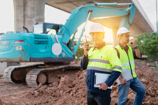 Two male engineers, meticulously review data on a laptop at a dynamic construction site. A large excavator and concrete bridge structure form the industrial background