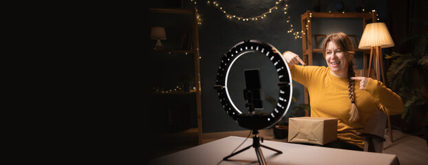 Fototapeta premium Female blogger unpacking box recording video using smartphone, ring light with tripod say Choose Me Pointing At Himself late evening at home office. Concept of social media