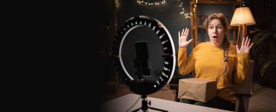 Female blogger unpacking box recording video using smartphone, ring light with tripod raising hands from sudden fear, showing stop, late evening at home office. Concept social media