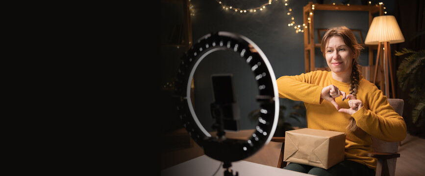 Female blogger unpacking box recording video using smartphone, ring light with tripod showing love heart gesture late evening at home office. Concept of online shopping and social media