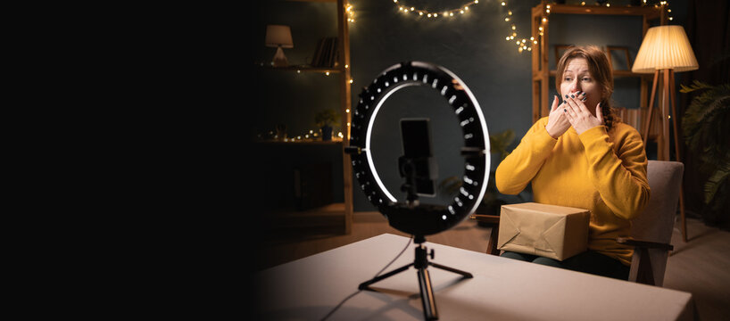 Shocked Female blogger unpacking box recording video using smartphone, ring light with tripod making announcement and omg gesture late evening at home office. social media